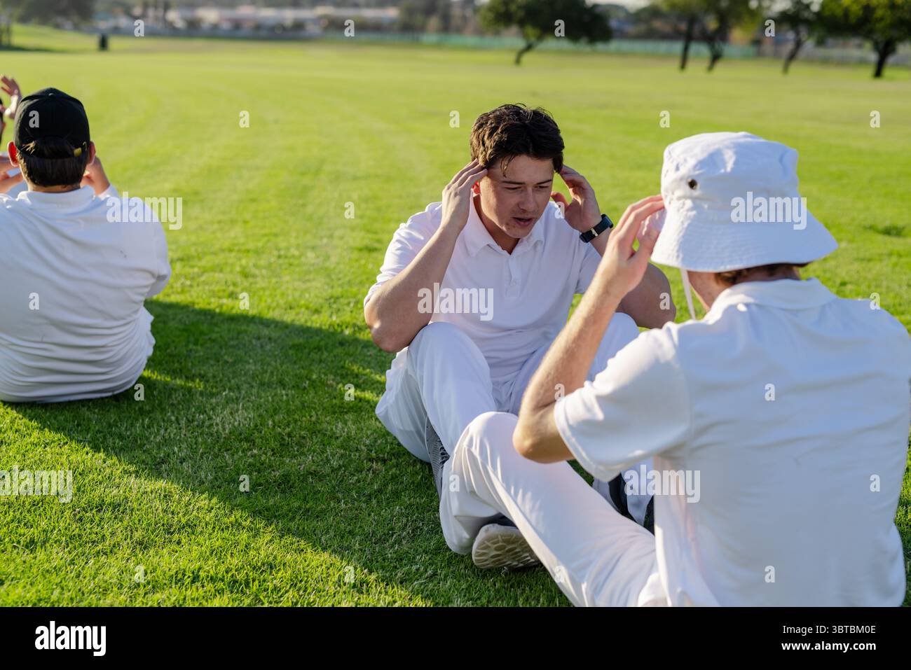 Compagni di squadra maschi seduti sul campo d'erba che eseguono esercizi da seduti con cappello da secchio e orologio da polso Foto Stock