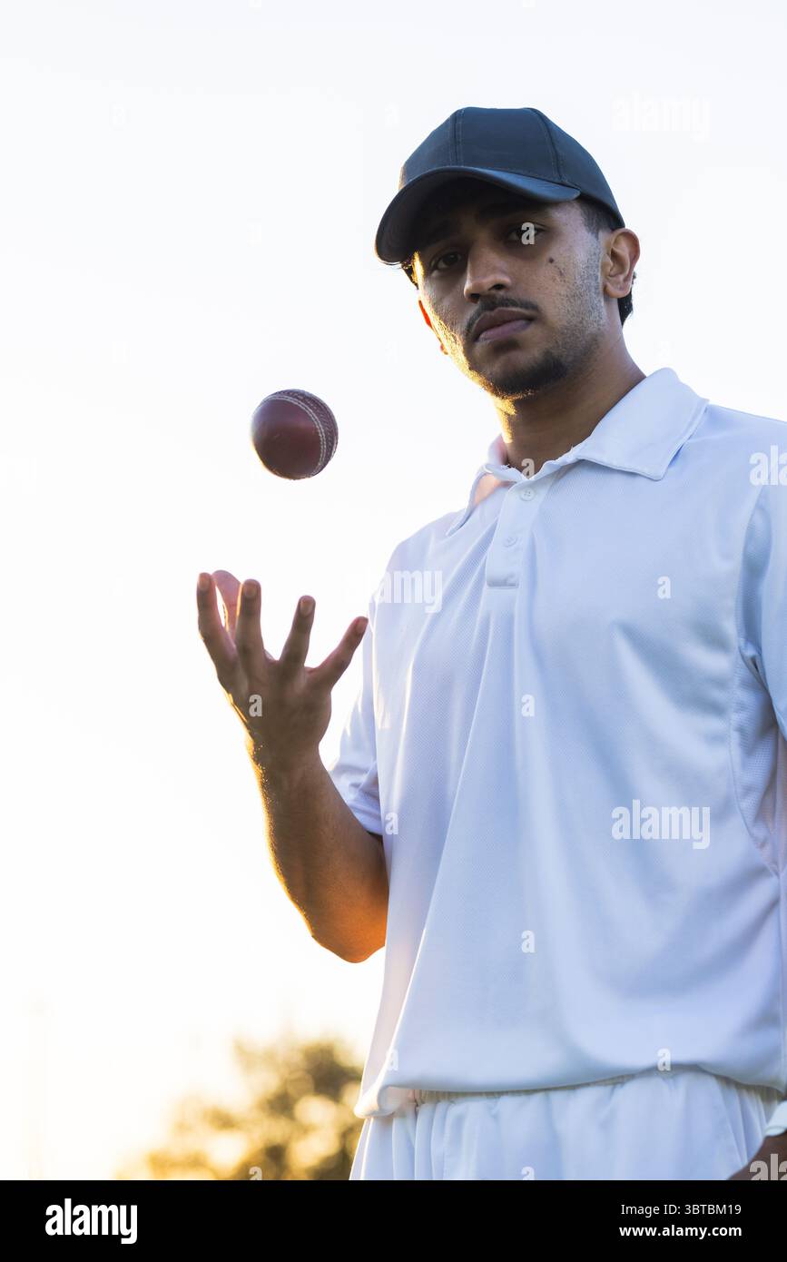 Uomo indiano che lancia la palla di cricket in mezzo al tramonto sul campo in uniforme con cappello, copia spazio Foto Stock