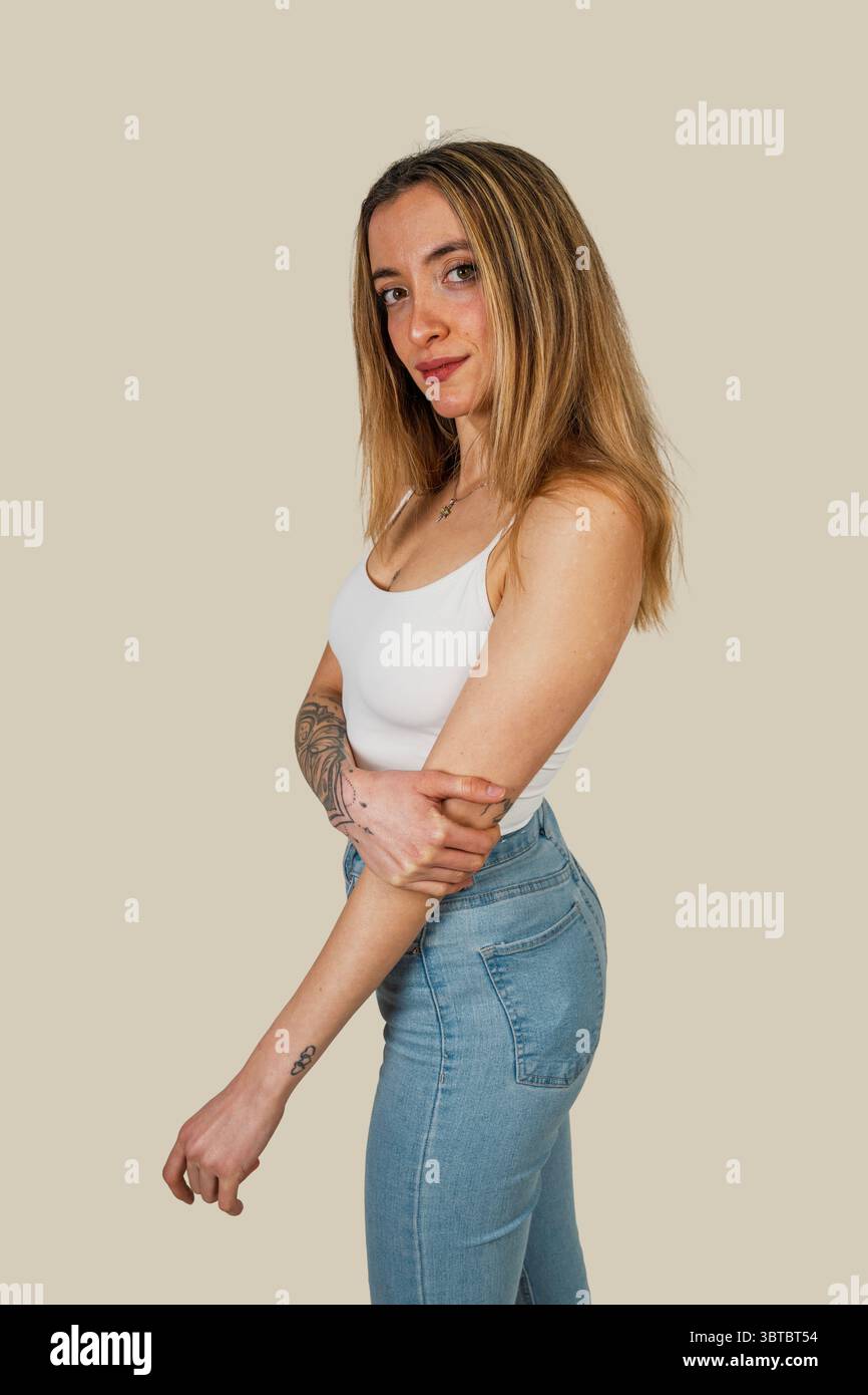 Ritratto in studio di una giovane donna tatuata che indossa un top bianco e jeans, in posa con le braccia incrociate su uno sfondo di colore chiaro Foto Stock