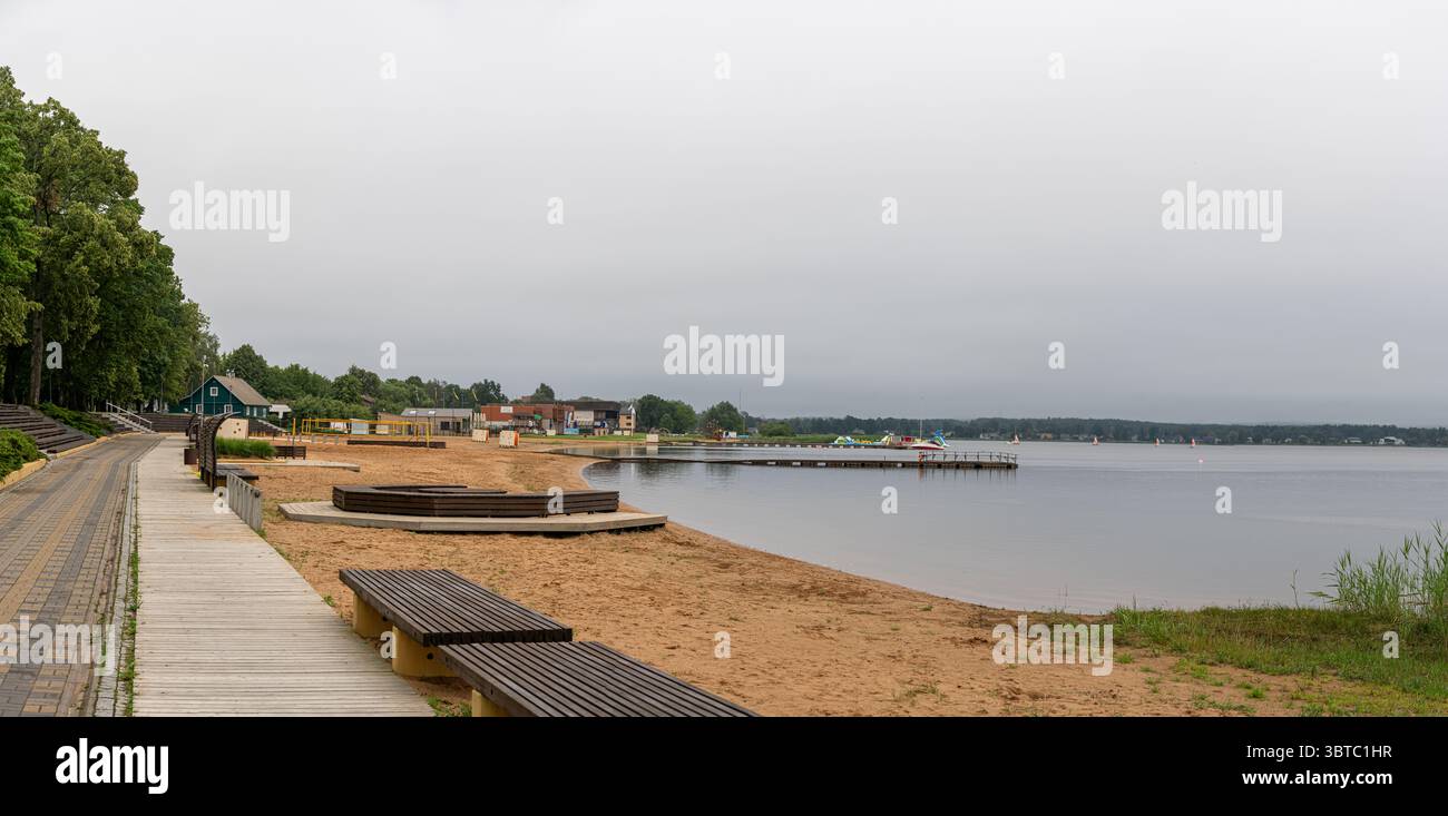 Vista panoramica sul lungomare del lago Tamula a Võru, Estonia: Spiaggia sabbiosa, passerella, panchine e molo sotto un cielo estivo nuvoloso. Foto Stock