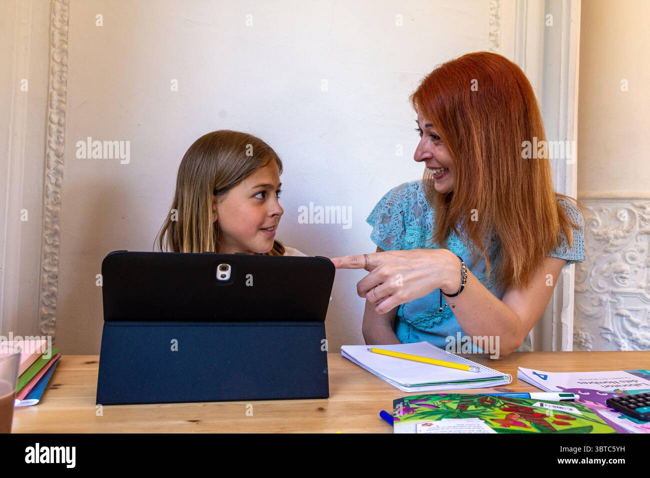 Madre rossa che assiste sua figlia bionda con i compiti scolastici utilizzando un tablet a casa, godendo del tempo di qualità insieme in un set di famiglia monoparente Foto Stock