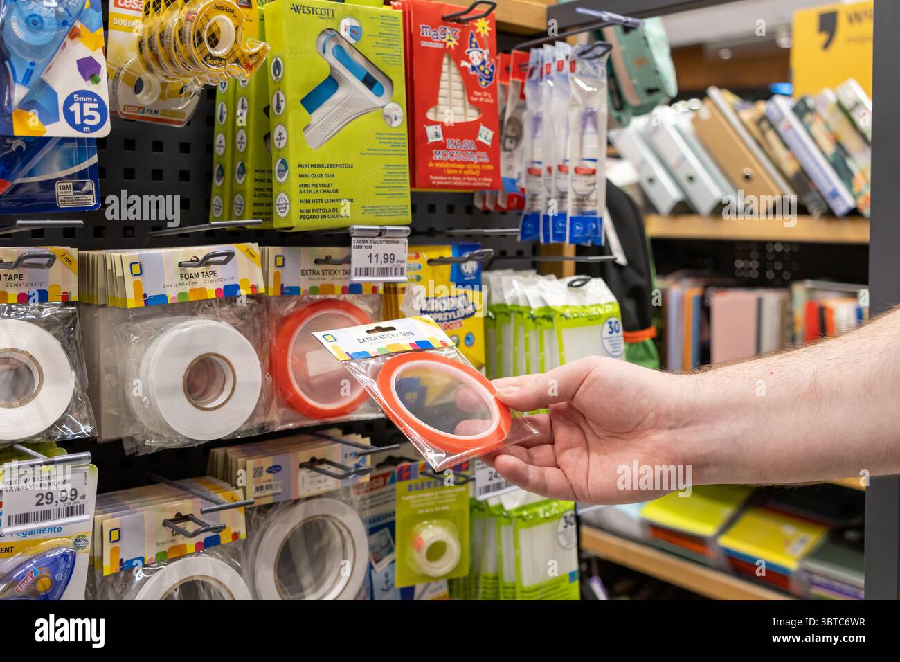 Poznan, Polonia - 30 giugno 2025: Un primo piano della mano di una persona che sceglie il nastro adesivo rosso da un espositore al dettaglio, con vari fornitori scolastici e di uffici Foto Stock