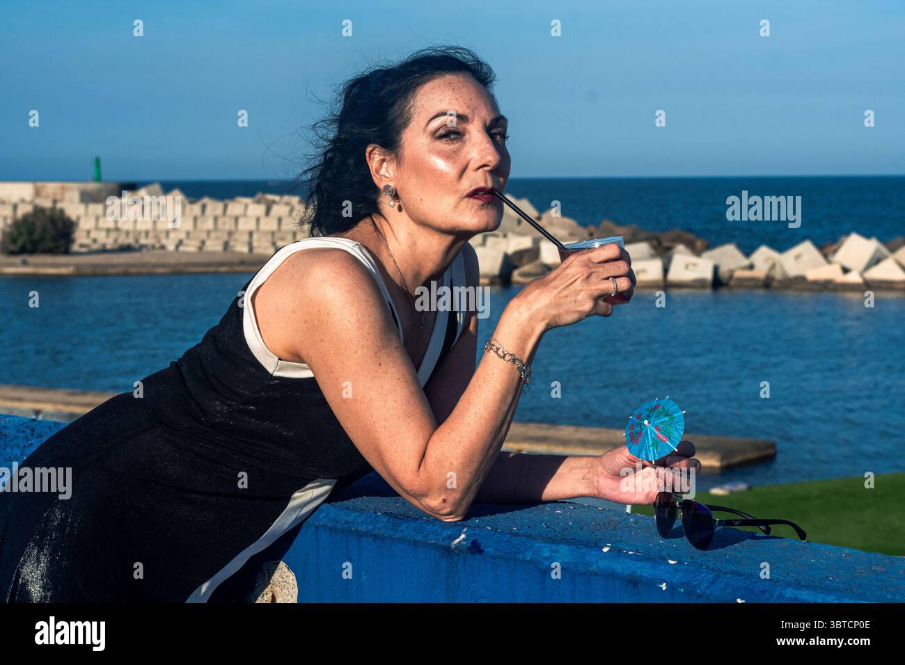 Elegante modello senior che assapora una bevanda vibrante sotto il sole caldo, irradia la positività del corpo e l'inclusione nel tranquillo mare blu, catturando l'ess Foto Stock