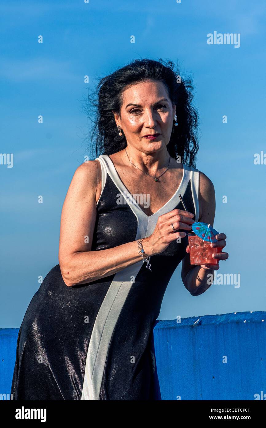 Elegante modello senior che abbraccia la positività e l'inclusione del corpo, in piedi all'aperto contro un cielo blu vibrante, tenendo in mano un cocktail con un ombrello in miniatura Foto Stock