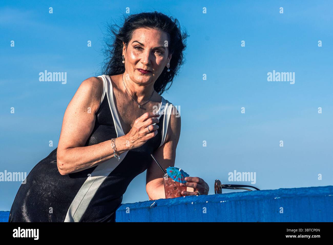 L'elegante modella senior gode di un cocktail rinfrescante in riva al mare in una giornata ventosa, i suoi capelli scorrono dolcemente nella brezza mentre assaporano il momento Foto Stock