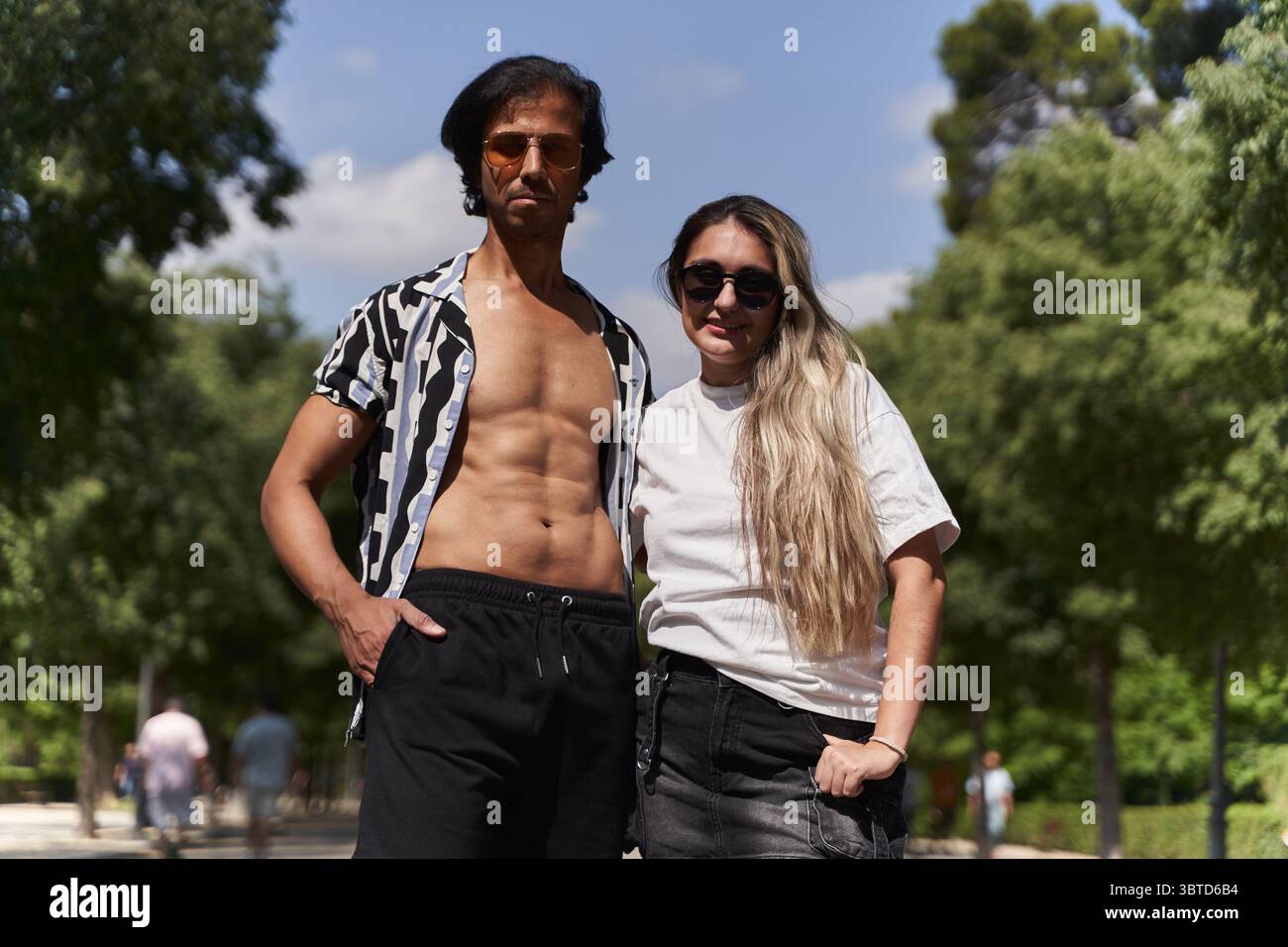 Coppia elegante che gode del sole estivo in un parco urbano, sfoggiando moda alla moda mentre si rilassa tra il verde e gli alberi Foto Stock