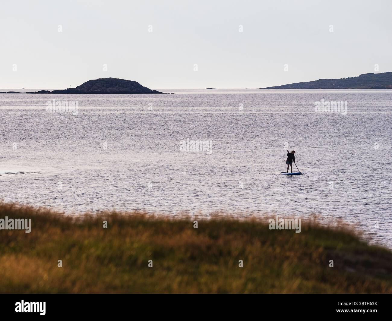 Una pedalatrice solitaria si muove senza sforzo attraverso acque tranquille, circondate da isole lontane sotto una luce soffusa e sbiadita. La scena riflette la B. Foto Stock