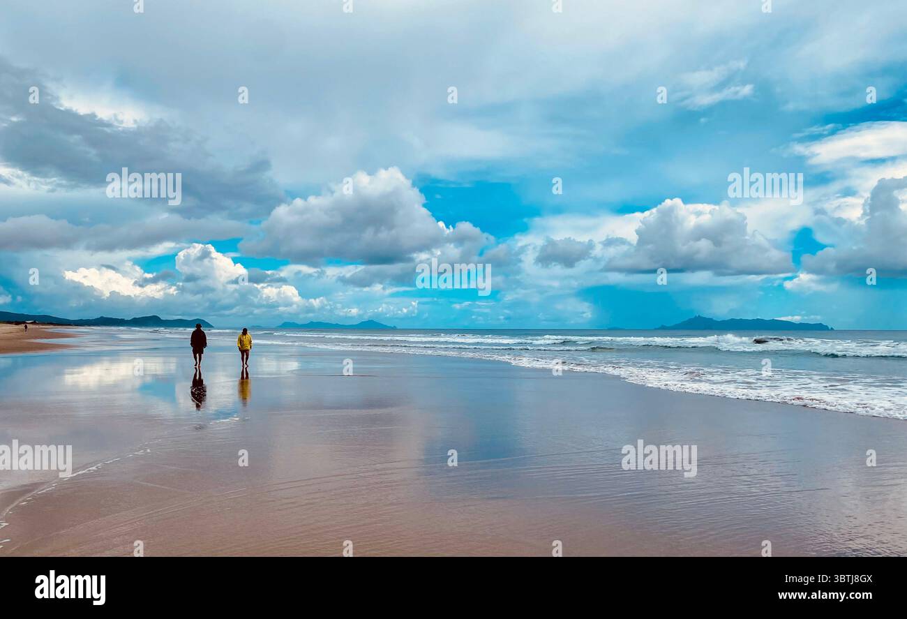 Due persone camminano su una spiaggia riflettente sotto un cielo blu con nuvole, con onde oceaniche e montagne lontane sullo sfondo. Foto Stock