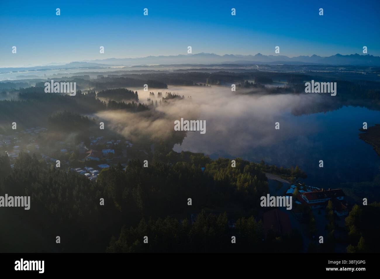 Vista aerea di un tranquillo lago abbracciato da una nebbia mistica, con le maestose Alpi in lontananza, Aitrang, Baviera, Germania. Foto Stock