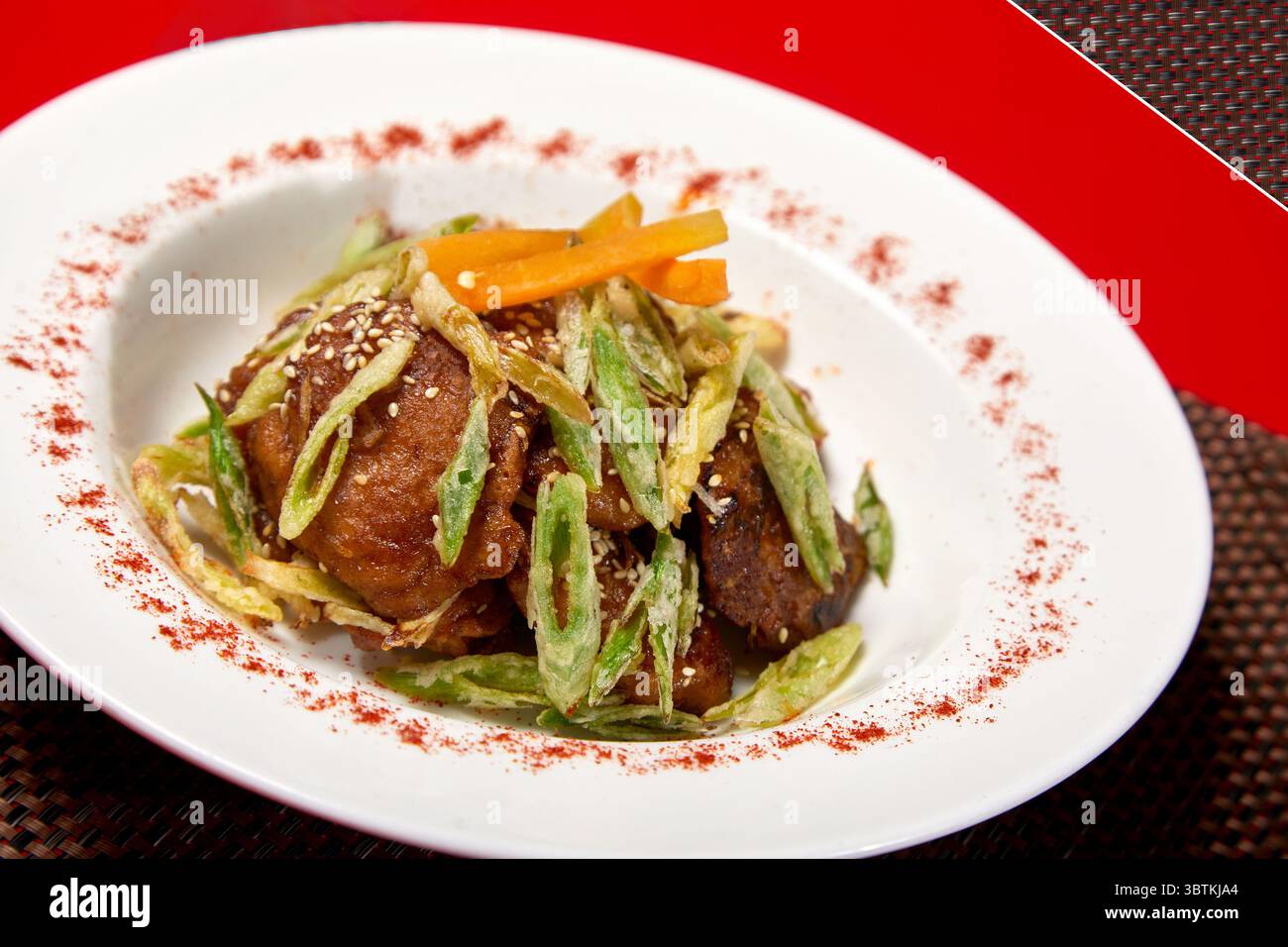 Deliziosi pezzi di pollo croccanti conditi con peperoncino verde fritto, carote julienate e semi di sesamo. Presentato su un piatto bianco con polvere di spezie rossa per Foto Stock