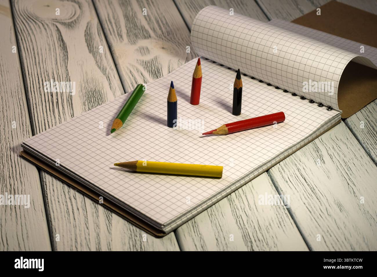 Blocco note vuoto aperto con carta a scacchi con matite colorate su un tavolo in legno bianco con sfondo didattico di cancelleria a luce crepuscolare Foto Stock