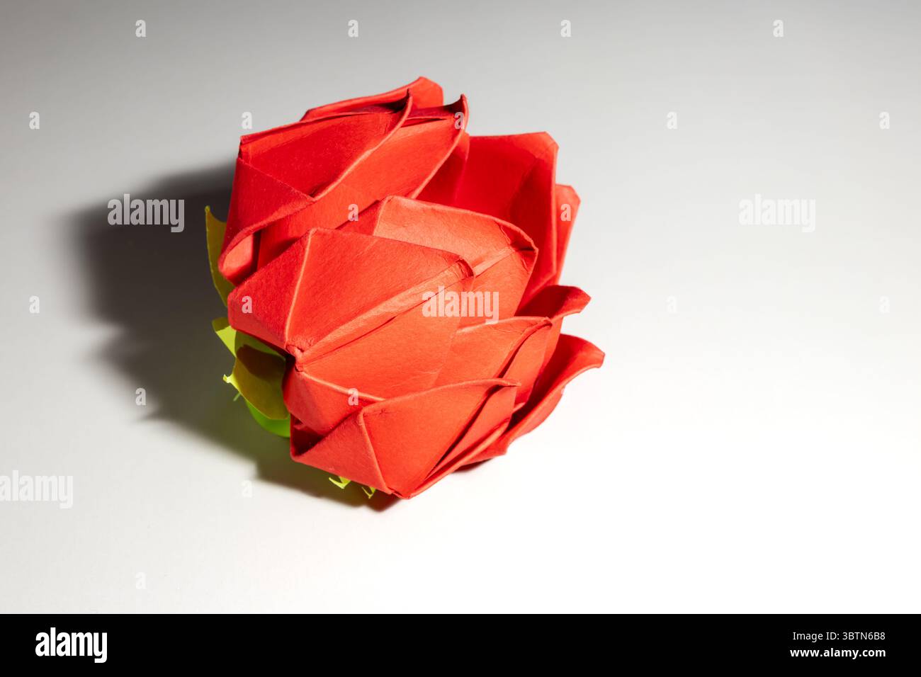 bocciolo di rosa rosso origami realizzato a mano con accenti verdi che creano ombre morbide su una superficie bianca pulita, sfoggiando intricate piegature di carta e delicate opere d'arte Foto Stock