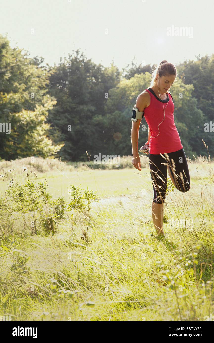 Runner femminile con cuffie che allunga la gamba in un parco soleggiato e erboso Foto Stock