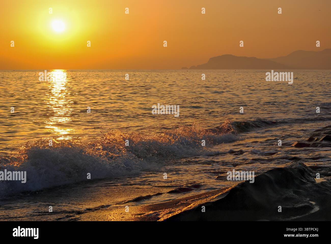 Tramonto dorato sul mare con dolci onde che colpiscono la riva e montagne lontane all'orizzonte. Foto Stock