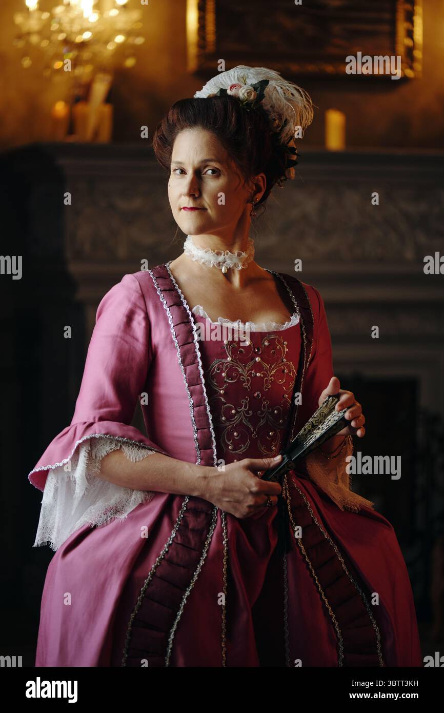 Ritratto di donna caucasica di mezza età che indossa un abito decorato in stile barocco che tiene in piedi un ventilatore decorativo in un ambiente interno storico ispirato al periodo rinascimentale Foto Stock