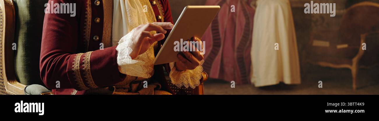 Uomo di mezza età caucasico che indossa un costume in stile barocco su un tablet digitale mentre è seduto all'interno, sfondo che mostra una figura sfocata in abiti rinascimentali, mescolando elementi storici e moderni Foto Stock