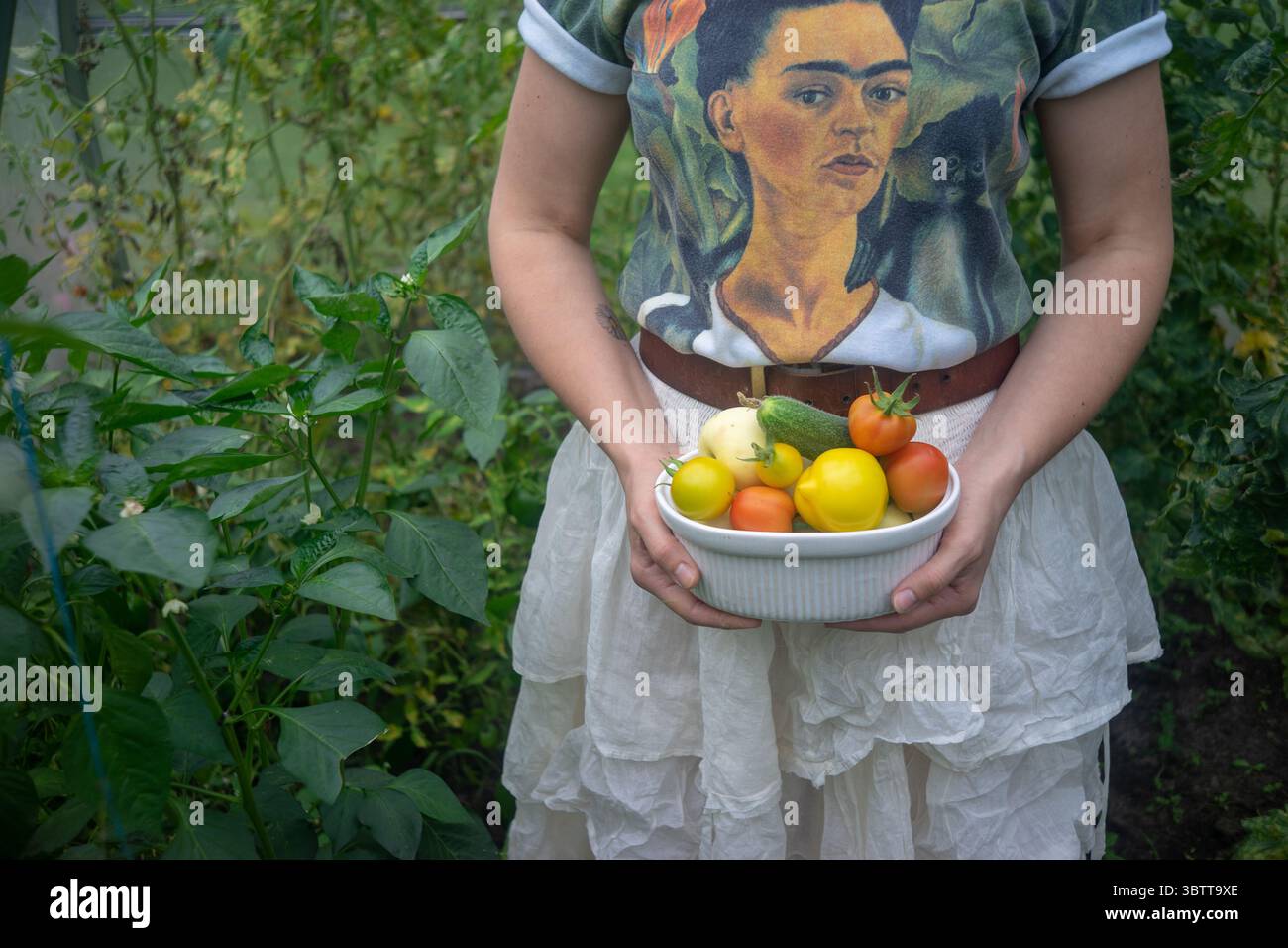 Una persona in piedi in un giardino lussureggiante con una ciotola bianca piena di verdure raccolte: Pomodori gialli e rossi, cetrioli Foto Stock