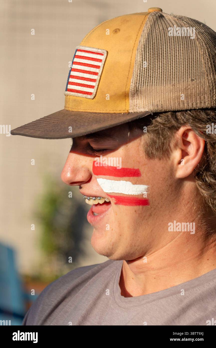 Un uomo indossa un cappello mentre mostra strisce rosse e bianche dipinte sul viso, creando un aspetto visivo sorprendente Foto Stock