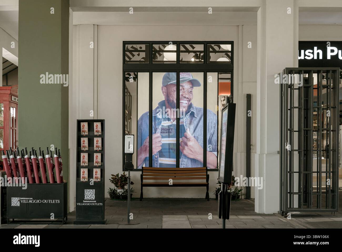 Karawang, Indonesia, 16 luglio 2025: Scena d'ingresso al Villaggio Outlets con una grande pubblicità di moda, una panchina vuota e un igienizzatore per le mani. Foto Stock