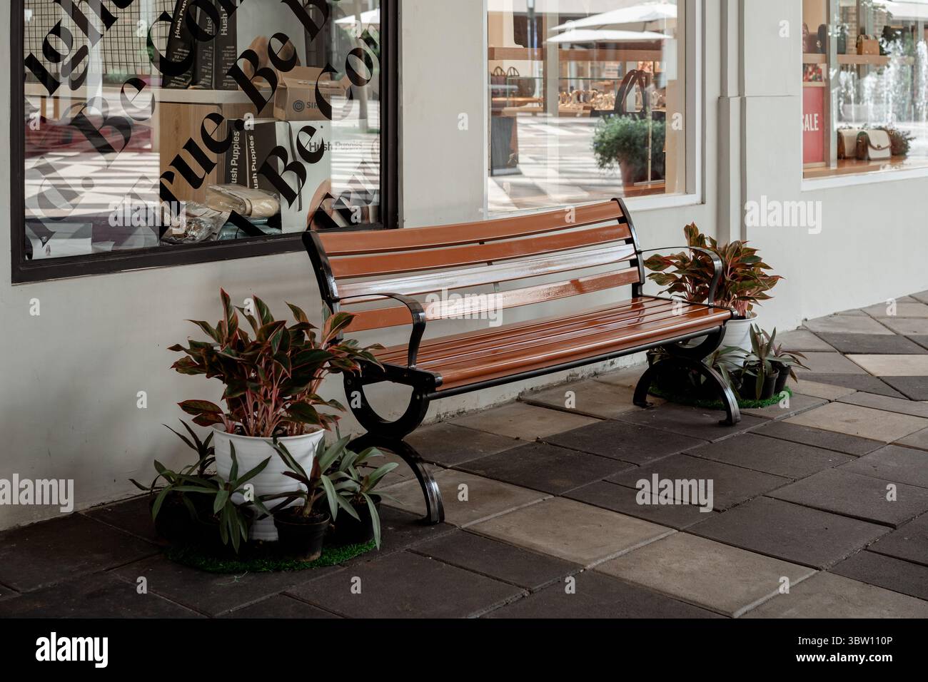 Karawang, Indonesia, 16 luglio 2025: Panca vuota del parco su un marciapiede piastrellato fuori dalla vetrina di un negozio, decorata con piante in vaso di Aglaonema. Foto Stock