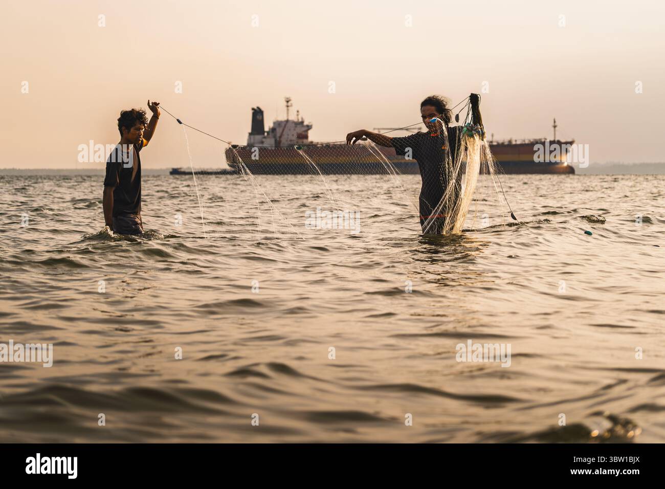 Balikpapan, Indonesia - 6 maggio 2025. Due pescatori locali gettano reti in acque costiere poco profonde con navi da carico sullo sfondo durante il Golden hou Foto Stock
