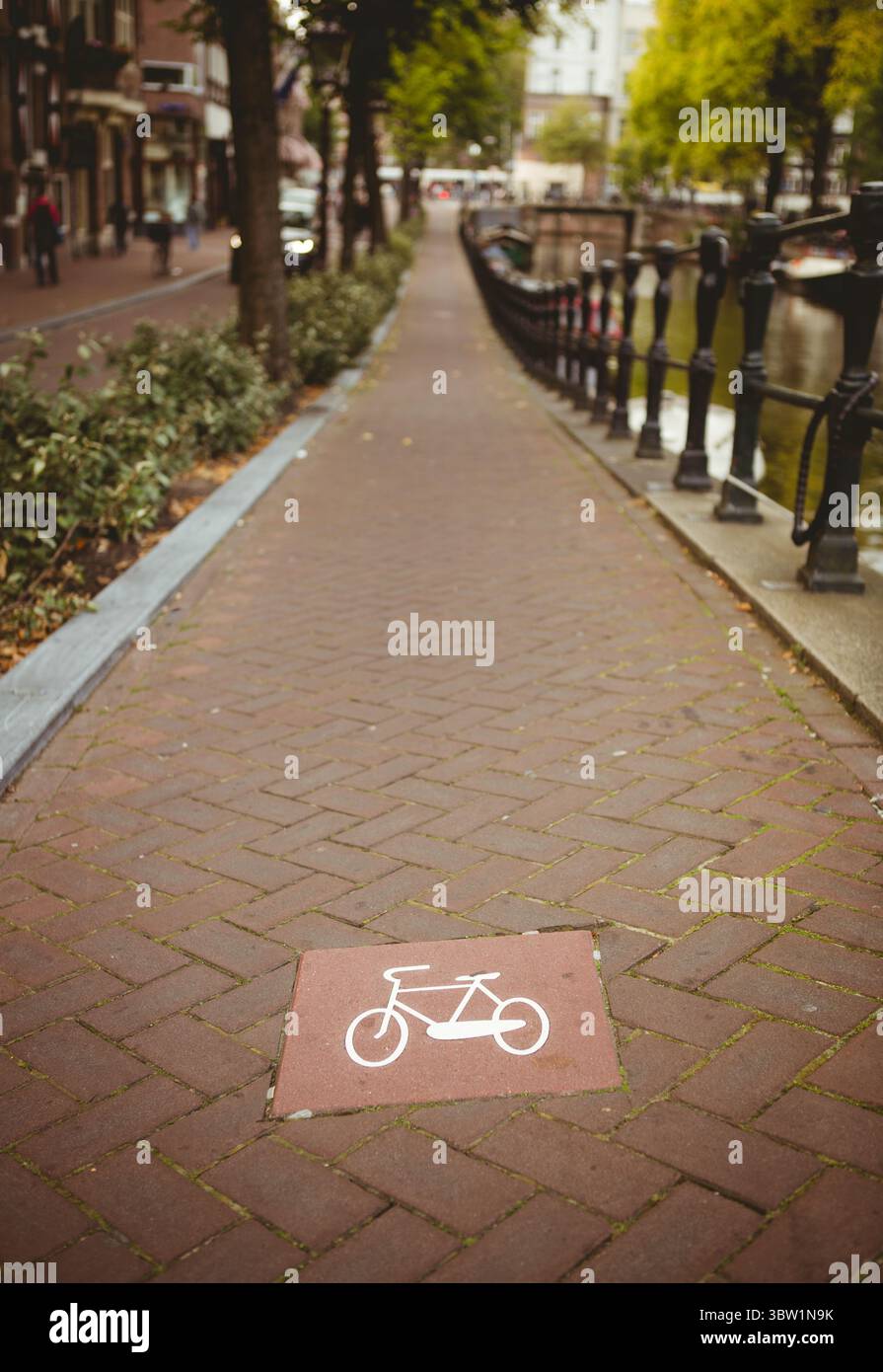 La piastrella rossa che mostra l'icona della bicicletta sta segnando una pista ciclabile dal design piatto lungo il canale in mattoni con ringhiera Foto Stock