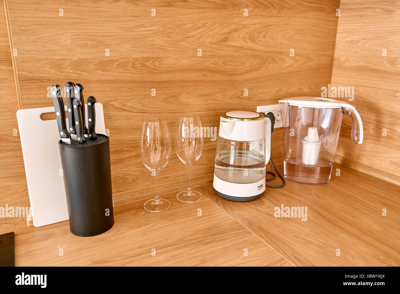 Un moderno piano di lavoro della cucina con una macchina per il caffè, set di coltelli, bicchieri da vino e una caraffa per filtro dell'acqua, il tutto sistemato in modo ordinato su una superficie in legno. Foto Stock