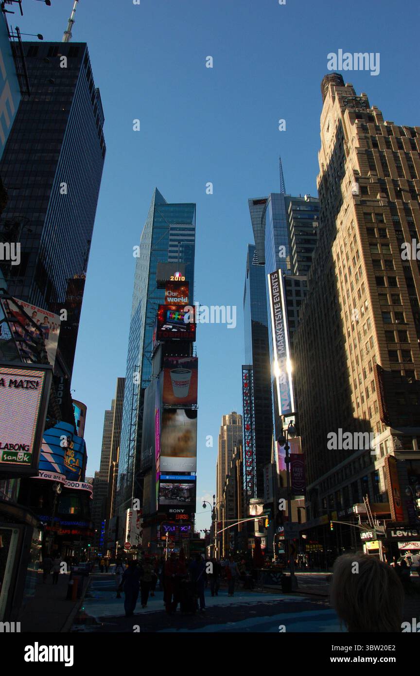 Times Square a New York City nel tardo pomeriggio, con grattacieli alti, cartelloni e persone che camminano sotto un cielo azzurro Foto Stock