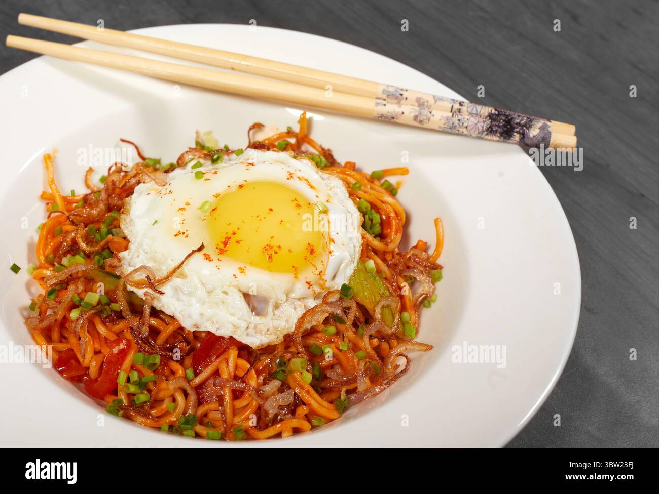 Deliziosi spaghetti fritti speziati con un uovo con il lato soleggiato, guarniti con scalogni e scalogni croccanti. Perfettamente placcato con bacchette e flor Foto Stock