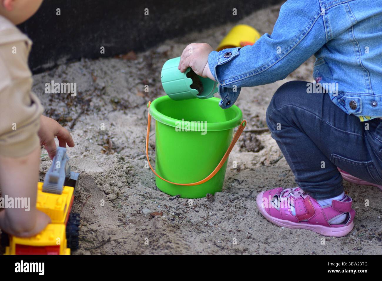 Due bambini piccoli che giocano in una scatola di sabbia con secchio verde e camion giocattolo. Primo piano di mani per bambini e giocattoli colorati, che esprimono divertimento all'aperto, creatività, Foto Stock
