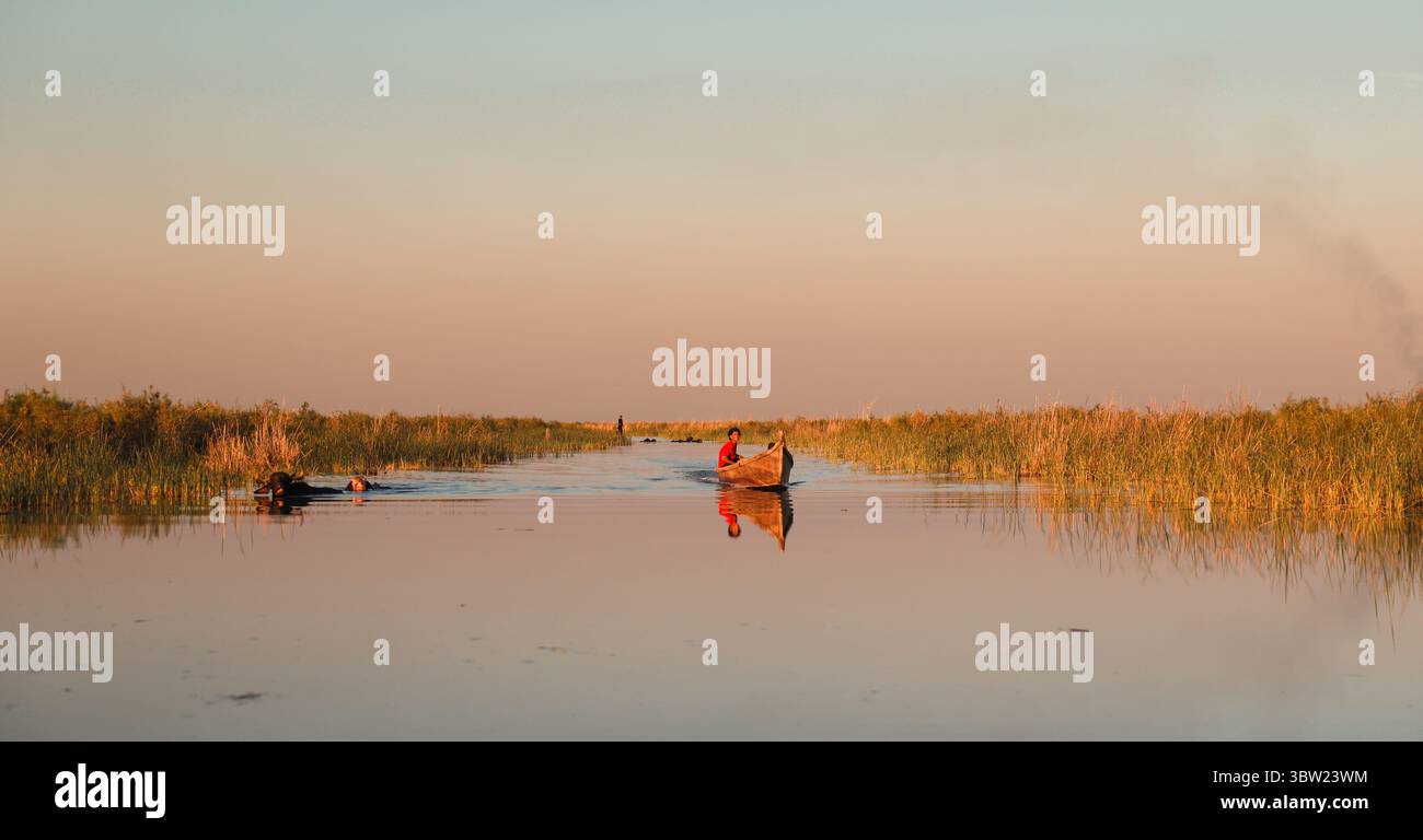 Un tranquillo corso d'acqua nelle paludi della Mesopotamica mostra gli abitanti locali su barche tradizionali durante il tramonto, circondati da lussureggianti canne. Foto Stock