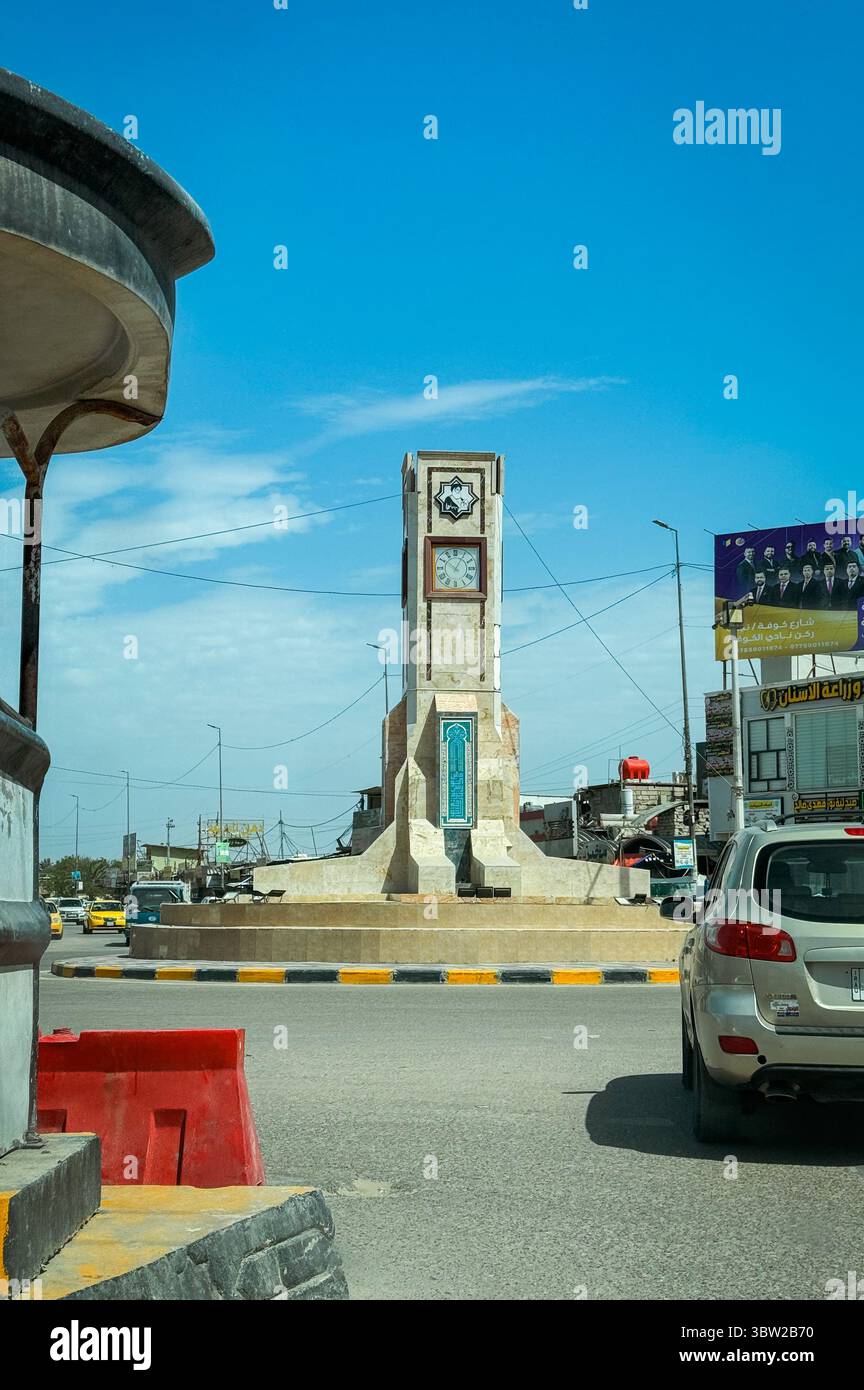 Una torre dell'orologio funge da punto focale in un trafficato incrocio di Baghdad, circondato da veicoli e cieli azzurri. Foto Stock
