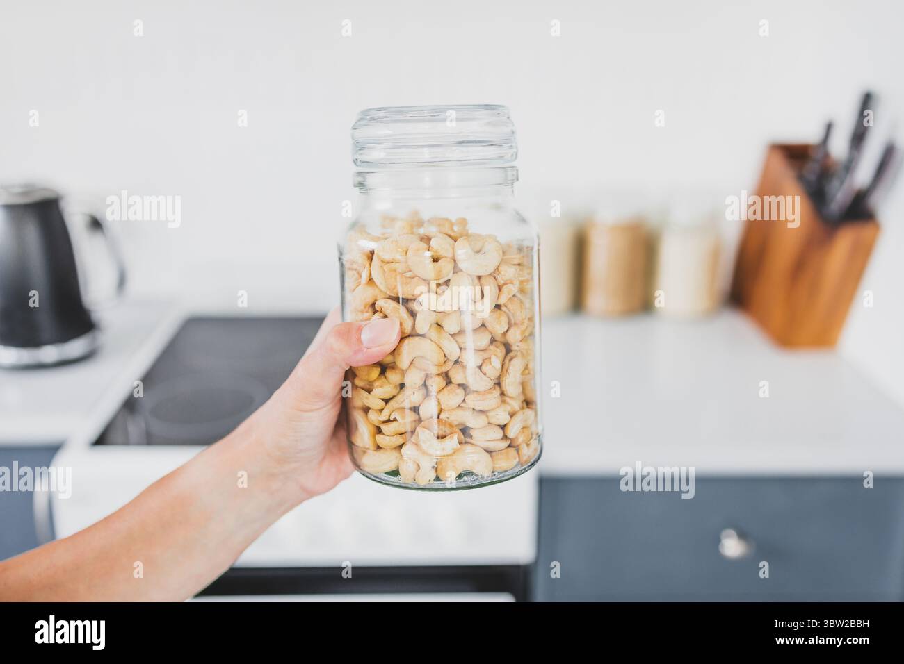 tenendo a mano un vaso di vetro di anacardi di fronte al bokeh della cucina, concetto di cucinare da zero e avere una dispensa organizzata Foto Stock tenendo a mano un vaso di vetro di anacardi di fronte al bokeh della cucina, concetto di cucinare da zero e avere una dispensa organizzata Foto Stock