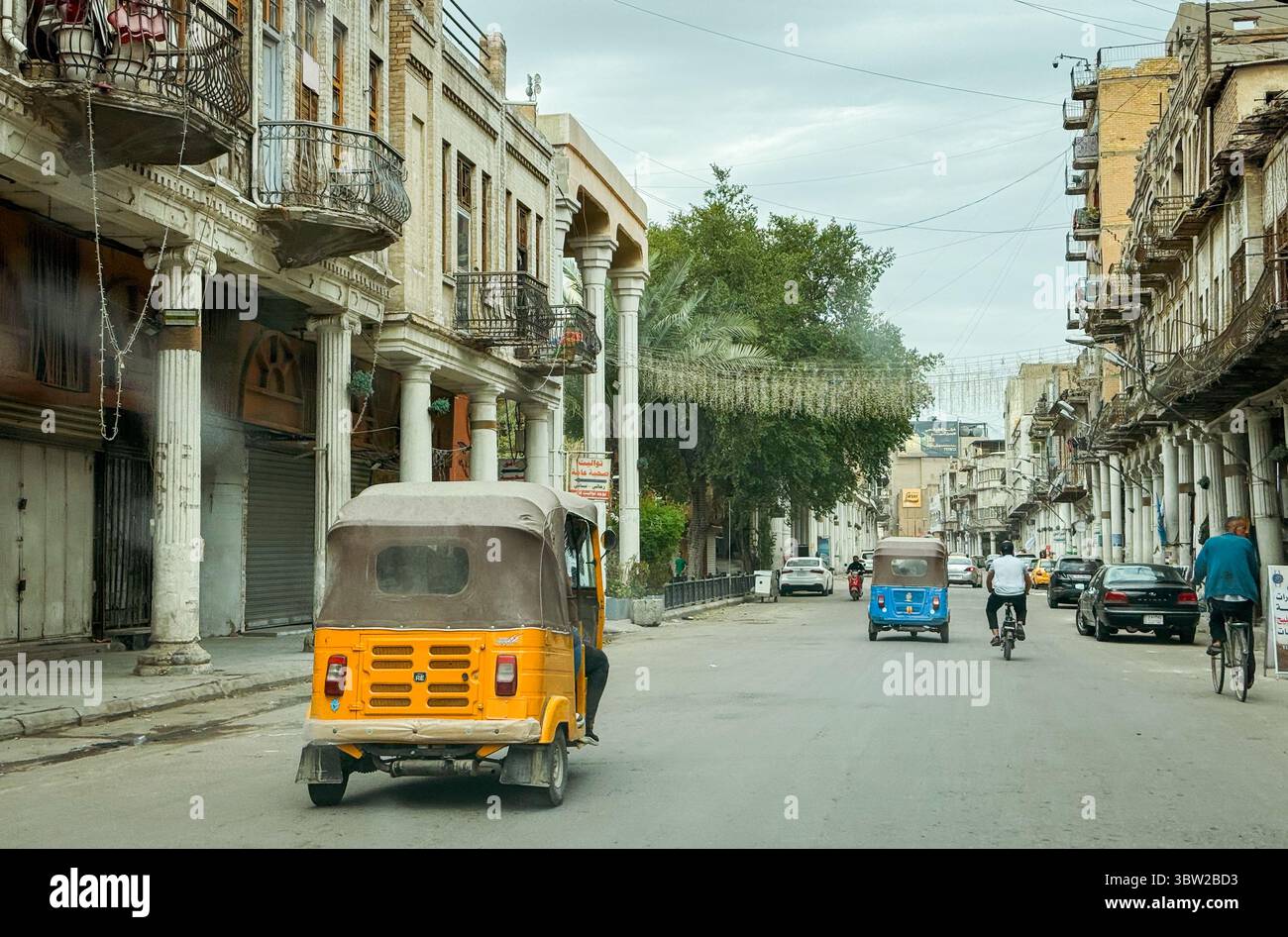 Tuk tuk e ciclisti navigano in una vivace strada di Baghdad, mostrando l'attività quotidiana e la vita urbana della città. Foto Stock