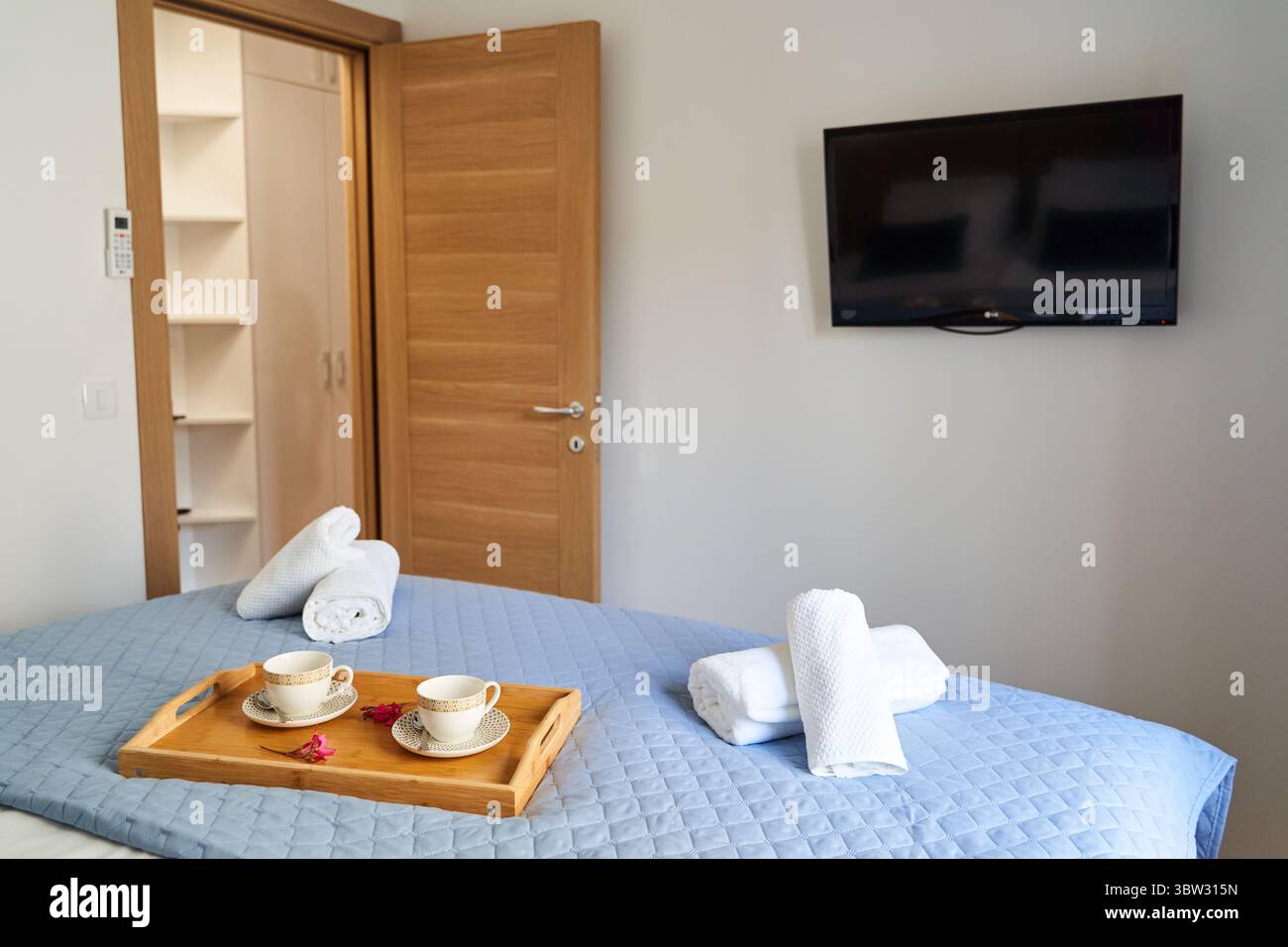 Un'intima camera da letto presenta un letto ben fatto con un vassoio di ristoro, una tv a parete e una porta aperta in legno che conduce ad un armadio. lo spazio trasuda tr Foto Stock