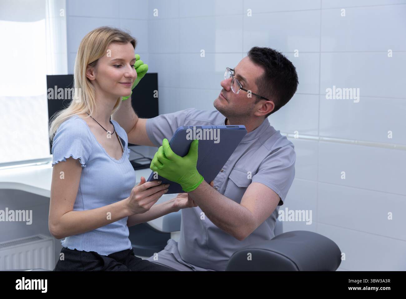 Una consulenza professionale in una clinica estetica moderna: Esperto che valuta le caratteristiche del viso e la salute della pelle del cliente con tablet per personalizzare Foto Stock