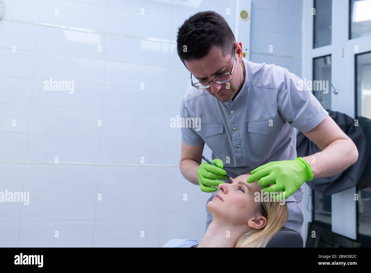 Un estetista professionista che esegue un trattamento cosmetico di precisione su un paziente in un ambiente clinico moderno con pareti piastrellate bianche pulite e luminose Foto Stock