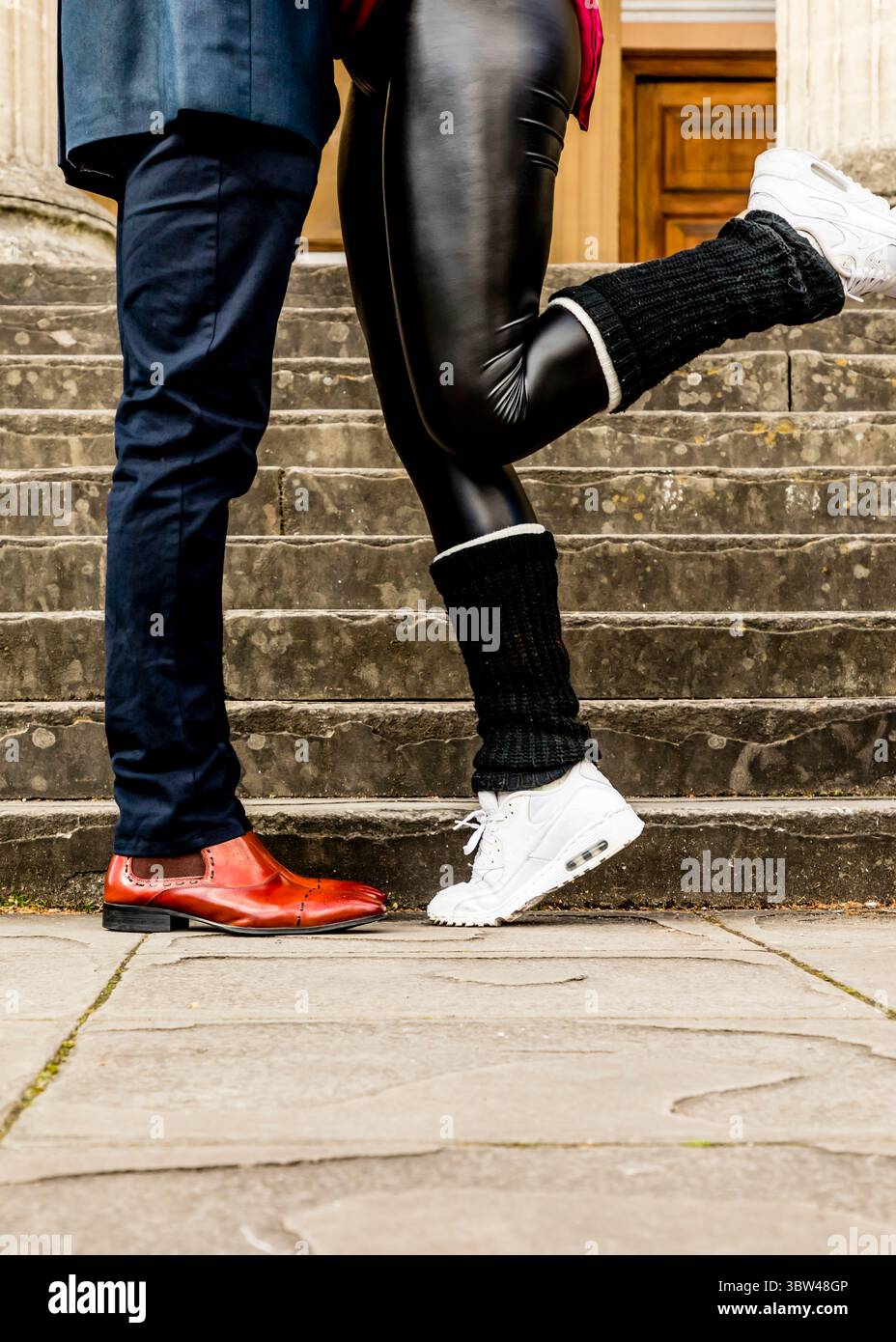 Un uomo con un vestito e scarpe rosse si trova di fronte a una persona che indossa leggings neri, sneakers bianche e scalda gambe. Foto Stock