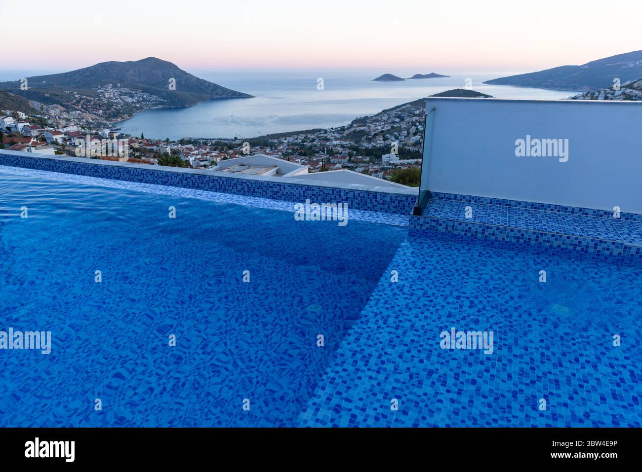 Tranquilla piscina a sfioro con una tranquilla vista sul mare di una città costiera al tramonto, circondata da colline Foto Stock Tranquilla piscina a sfioro con una tranquilla vista sul mare di una città costiera al tramonto, circondata da colline Foto Stock