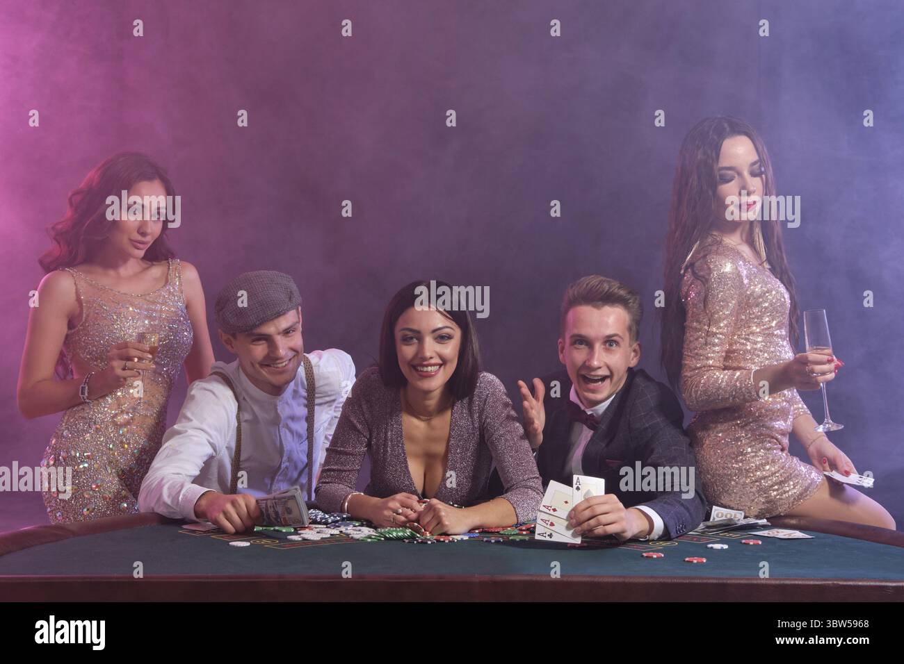 I colleghi a giocare a poker al Casino, a tavola con pile di chips, denaro, carte su di esso. Felici di vincere, bevendo champagne, sorridente. Nero fumo, backg Foto Stock