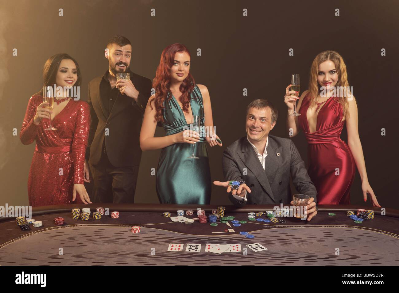 I colleghi felici giocano a poker al casinò. Festeggiano la vittoria, sorridono e sembrano eccitati mentre posano al tavolo contro un buio Foto Stock