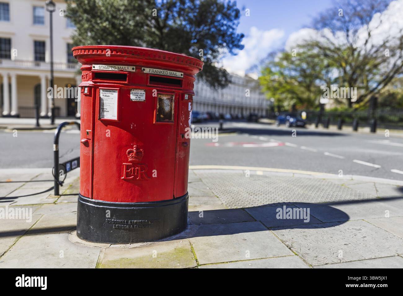 Primo piano di una tradizionale cassetta postale rossa britannica su un marciapiede della città con cieli blu e alberi frondosi alle spalle. Londra, Regno Unito, 1° aprile 2024 Foto Stock