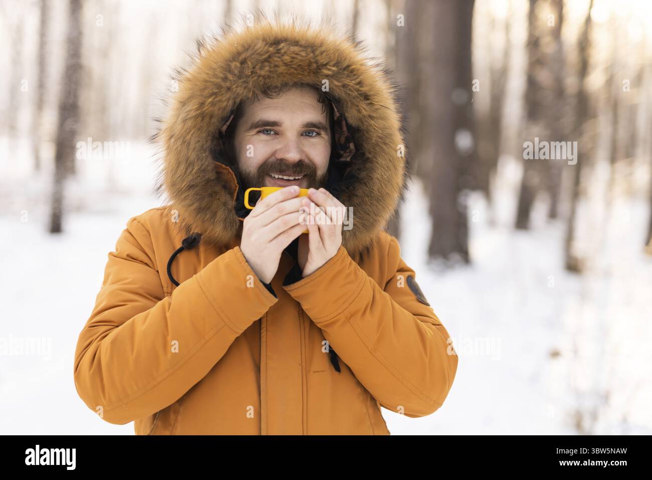 Uomo con tazza gialla nella foresta innevata che guarda la macchina fotografica. Consapevolezza, calore e connessione umana con la natura durante una tranquilla pausa invernale. Servizio fotocopie Foto Stock