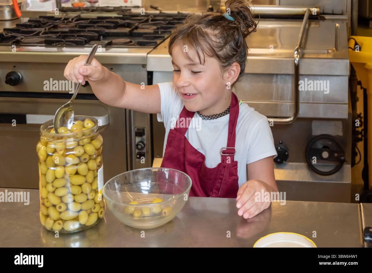 Una ragazza in cucina in un grembiule tira le olive da un vaso grande usando un cucchiaio, preparandosi a cucinare un pasto Foto Stock