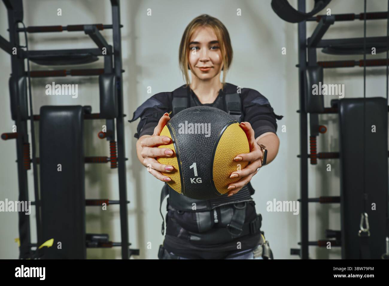Attraente femmina bionda che indossa nei sensori e cinghie di fissaggio con Velcro è la pratica di EMS fitness in una palestra, costruendo i muscoli con un Bal ponderata Foto Stock