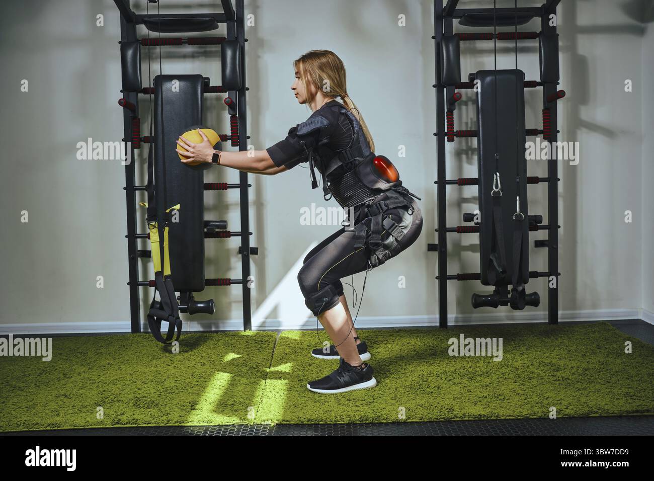 Una splendida bionda signora che indossa nei sensori e cinghie di fissaggio con Velcro è la pratica di EMS fitness in una palestra, costruire muscoli e facendo squat con un Foto Stock