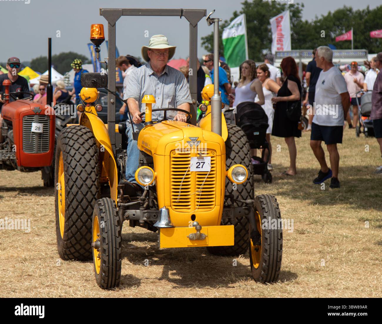 Un uomo con cappello di paglia che guida un trattore giallo Masset Ferguson 35 a una fiera agricola in una giornata di sole. Foto Stock