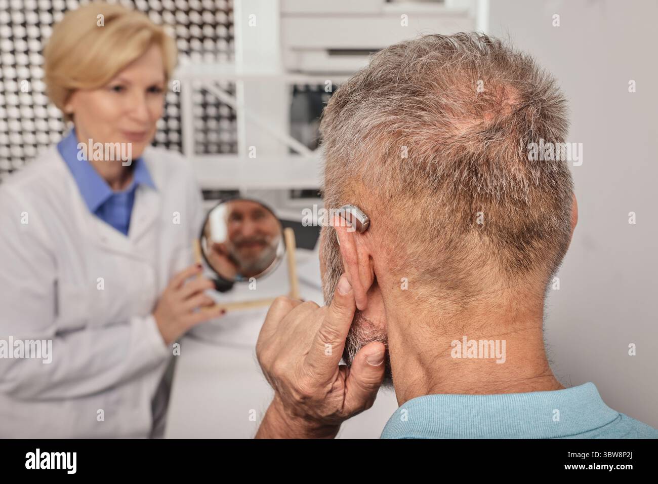 Uomo anziano che prova l'apparecchio acustico di fronte allo specchio con audiologa donna che mette l'apparecchio all'orecchio durante la consultazione audiologica Foto Stock