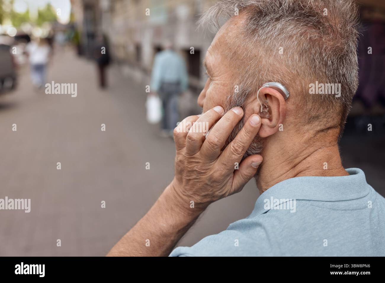 L'uomo anziano con l'apparecchio acustico dietro l'orecchio tiene la mano vicino all'orecchio per ascoltare l'ambiente urbano, avendo piena vita Foto Stock
