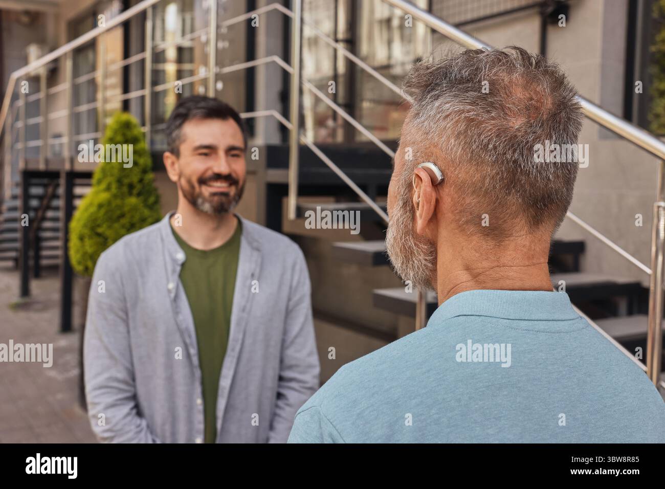 Soluzioni per l'udito. Un uomo anziano positivo con disturbi dell'udito utilizza un apparecchio acustico per comunicare con il figlio adulto in strada urbana Foto Stock